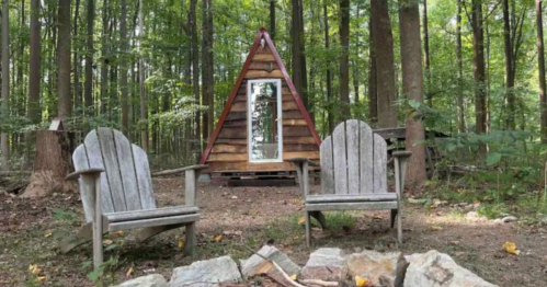A cozy A-frame cabin surrounded by trees, with two wooden chairs in the foreground and a fire pit nearby.