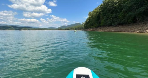 A paddleboarder glides across calm, green water with lush hills and a blue sky in the background.
