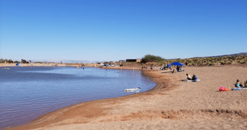 A sandy beach by a calm lake with people relaxing, swimming, and enjoying the sunny day under a clear blue sky.
