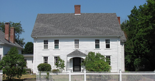 A large white two-story house with a gray roof, surrounded by greenery and a white picket fence.