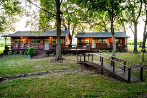 Two rustic cabins surrounded by trees, connected by a wooden bridge, set in a grassy area with soft evening light.