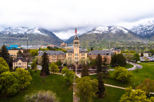 Aerial view of a university campus with a historic building, surrounded by trees and mountains under a cloudy sky.