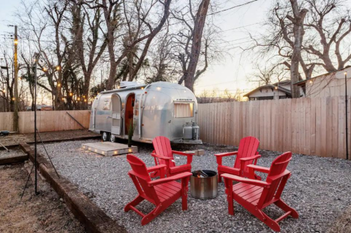 A vintage Airstream trailer in a backyard with red Adirondack chairs and a fire pit, surrounded by trees and a wooden fence.