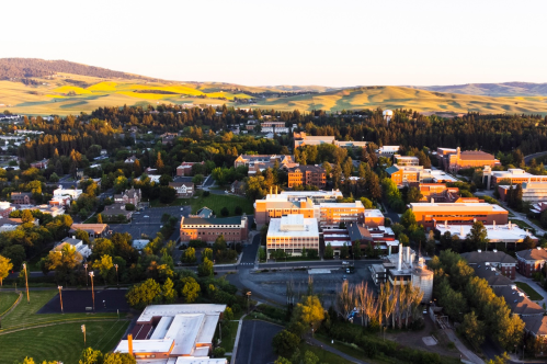 Aerial view of a university campus surrounded by green hills and trees, with buildings and sports fields visible.