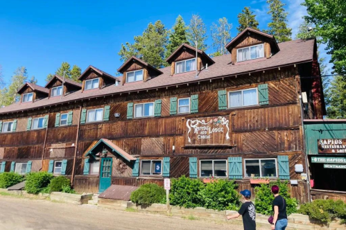 A rustic wooden lodge with multiple gabled roofs, surrounded by trees and a clear blue sky. Two people walk by.