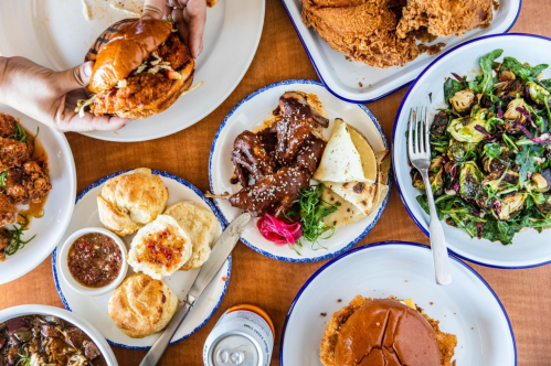 A table spread with fried chicken, sandwiches, salads, biscuits, and sauces, showcasing a variety of delicious dishes.