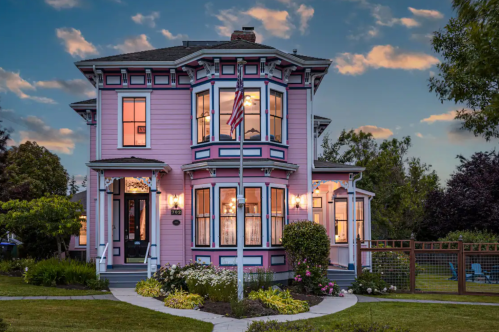 A charming pink Victorian house with decorative trim, surrounded by lush greenery and a flagpole, under a twilight sky.