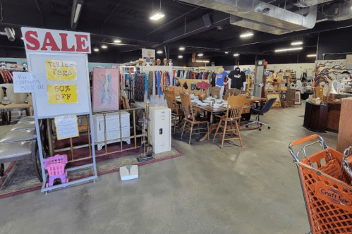 A spacious thrift store with sale signs, clothing racks, and tables set for dining, featuring an orange shopping cart.