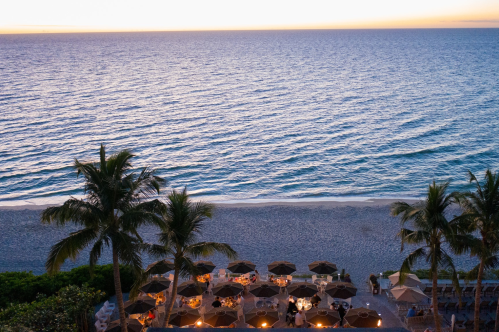 A serene beach at sunset, with palm trees and dining tables under umbrellas along the shore.