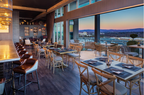 Modern restaurant interior with wooden tables, chairs, and a bar, featuring large windows and a scenic view at sunset.