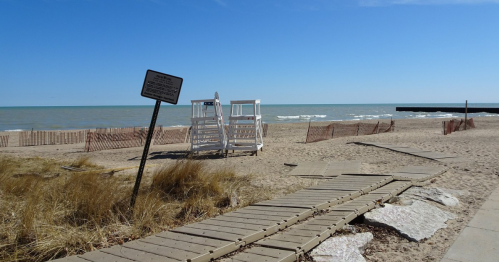 A sandy beach with a lifeguard stand, wooden walkway, and a sign, overlooking calm waves under a clear blue sky.