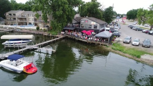 Aerial view of a busy lakeside restaurant with outdoor seating, boats docked, and cars parked nearby.