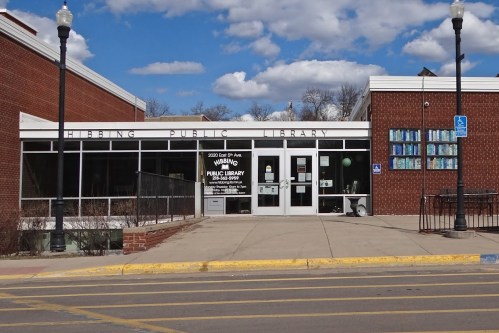 Hibbing Public Library entrance with large windows, a sign, and blue sky with clouds in the background.