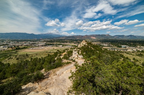 A panoramic view of a rocky ridge overlooking a valley and distant mountains under a partly cloudy sky.