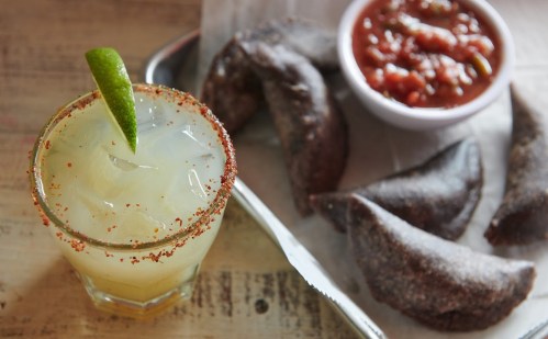 A refreshing cocktail with a lime wedge beside a plate of dark, crispy pastries and a bowl of salsa.