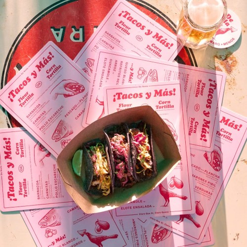 A tray of three tacos with colorful toppings, surrounded by menus and a glass of drink on a table.