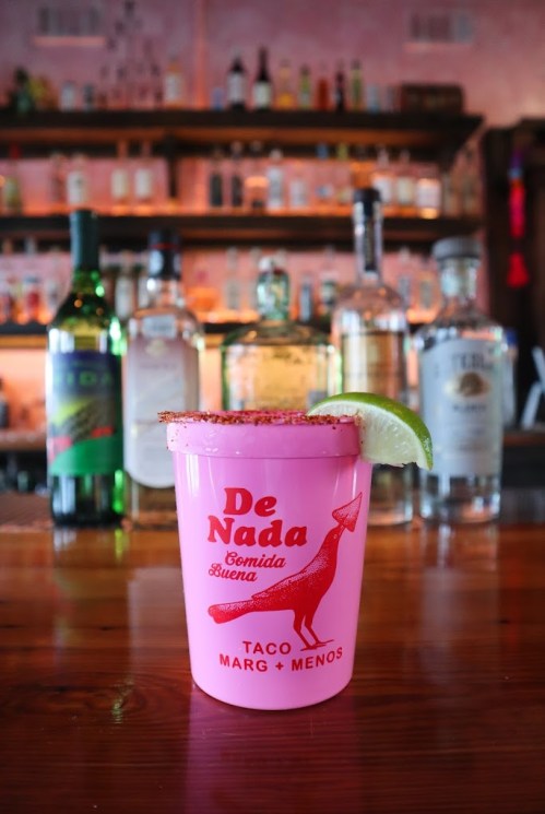 A pink cup with "De Nada" printed on it, filled with a margarita and garnished with a lime slice, on a wooden bar.
