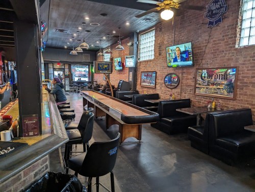 A spacious bar with a wooden counter, booths, and TVs on the walls, featuring a rustic brick interior.