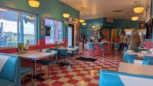 A colorful diner interior with checkered floors, teal and pink decor, and tables. People are seated and ordering at the counter.