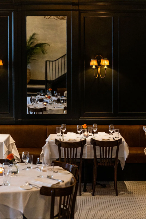 Elegant restaurant interior with white tablecloths, glassware, and a mirror reflecting a staircase and plants.