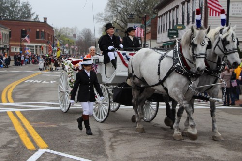 A horse-drawn carriage with riders in formal attire parades down a street, surrounded by flags and spectators.
