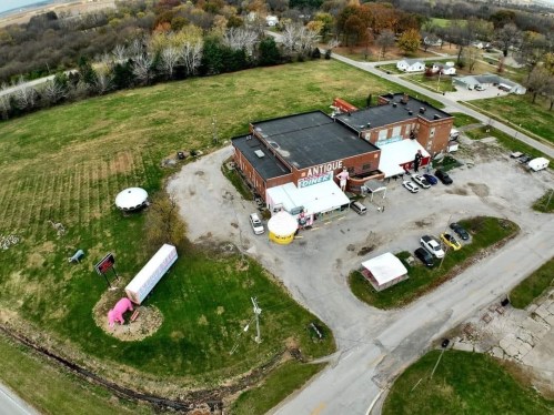 Aerial view of an antique store surrounded by fields and trees, with parked cars and a pink sculpture in the foreground.