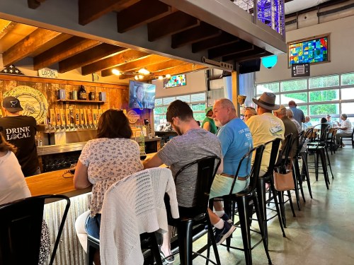 A bar scene with patrons sitting on stools, chatting, and enjoying drinks in a casual, well-lit setting.