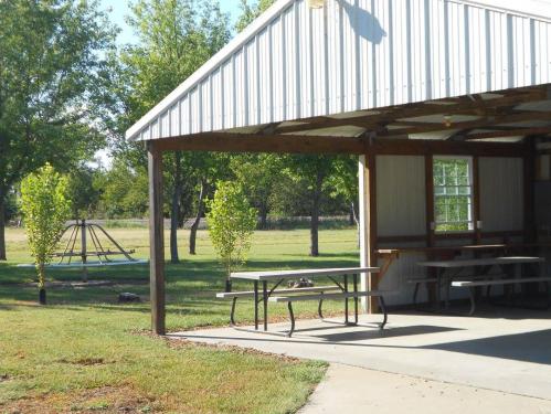 A shaded outdoor area with picnic tables, surrounded by green trees and a playground in the background.