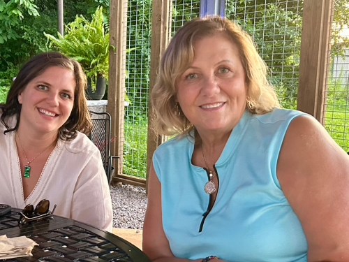 Two women smiling at a table outdoors, surrounded by greenery and a wooden structure.