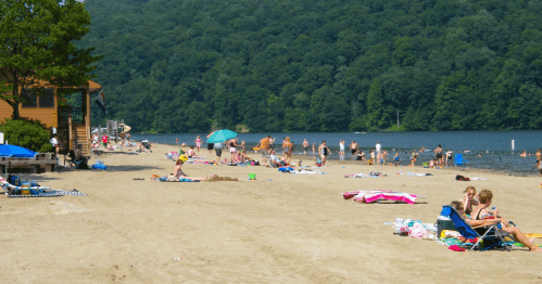 A sandy beach scene with people sunbathing, swimming, and relaxing near a lake, surrounded by green hills.