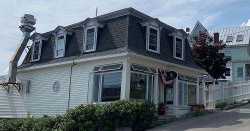 A charming two-story building with a sloped roof, featuring windows and decorative trim, surrounded by greenery.