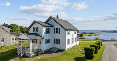 A white two-story house with a porch, overlooking a scenic waterfront and green landscape under a blue sky.
