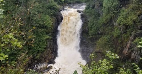 A powerful waterfall cascades down rocky cliffs, surrounded by lush green trees and a misty atmosphere.