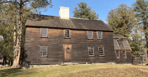 A historic wooden house with multiple windows, surrounded by trees and a clear blue sky.