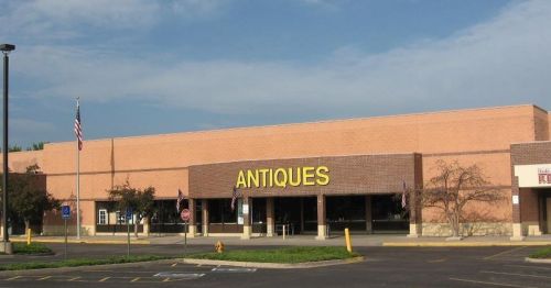 A brick building with a large sign reading "ANTIQUES," surrounded by a parking lot and blue sky.