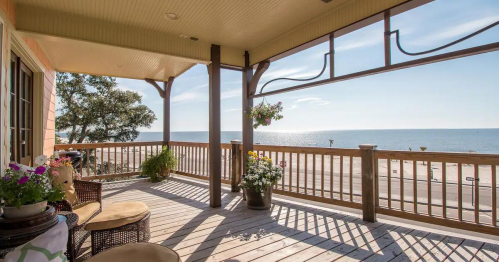 A sunny porch with seating and potted flowers overlooks a calm beach and sparkling ocean under a clear blue sky.