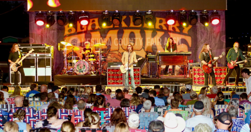 A band performs on stage at a fair, with a large audience seated in colorful chairs in front of them.