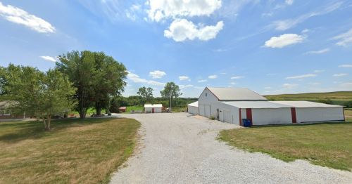 A gravel driveway leads to a farm with several buildings, trees, and a clear blue sky in the background.