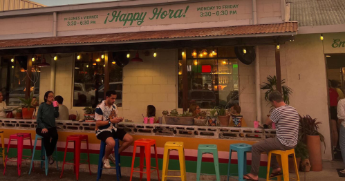 A colorful outdoor bar with patrons enjoying drinks, surrounded by plants and warm lighting, during happy hour.
