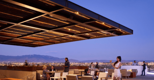 Rooftop bar with modern seating, people socializing, and a city skyline at dusk.