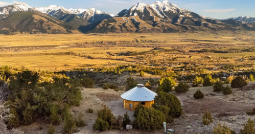 A cozy yurt nestled in a mountainous landscape, surrounded by trees and golden fields under a clear blue sky.