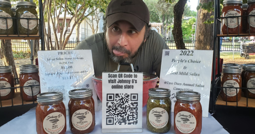 A vendor at a market displays jars of salsa with a QR code for online orders and signs for pricing and awards.