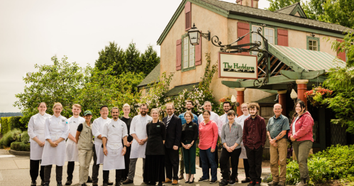A group of chefs and staff pose outside The Heron restaurant, surrounded by greenery and a charming building.