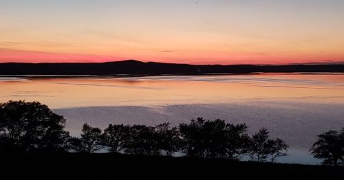 A serene sunset over a calm lake, with silhouettes of trees and distant hills reflecting vibrant colors in the water.