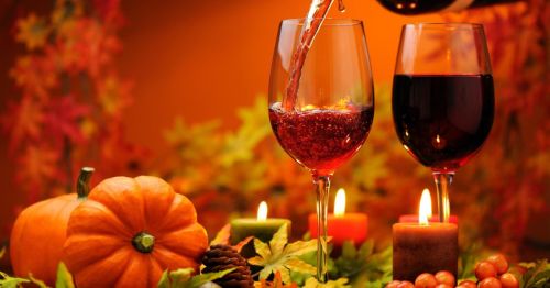 Two glasses of red wine are filled, surrounded by autumn leaves, a pumpkin, candles, and festive decorations.