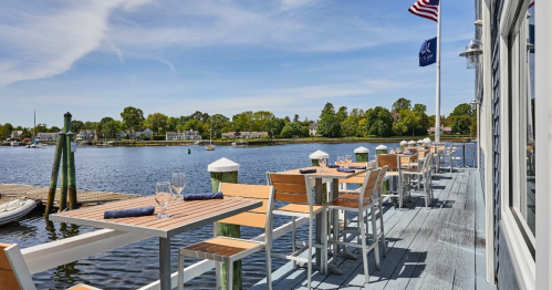 Outdoor dining area by a calm waterfront, featuring wooden tables and chairs, with a clear blue sky and American flag.