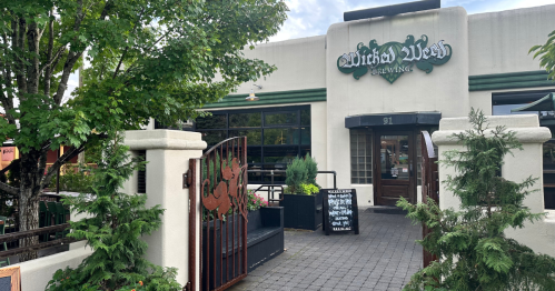 Exterior of Witch's Weed Brewing, featuring a green sign, entrance gate, and surrounding greenery.