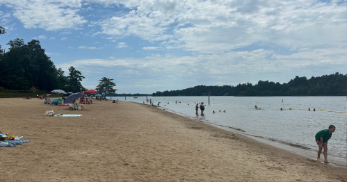 A sandy beach with people swimming and relaxing, surrounded by trees and a calm lake under a partly cloudy sky.