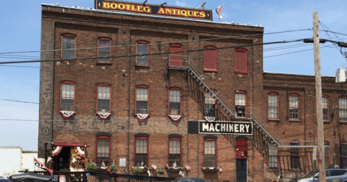 A historic brick building with red windows, featuring "Bootleg Antiques" and "Machinery" signs, and decorative bunting.