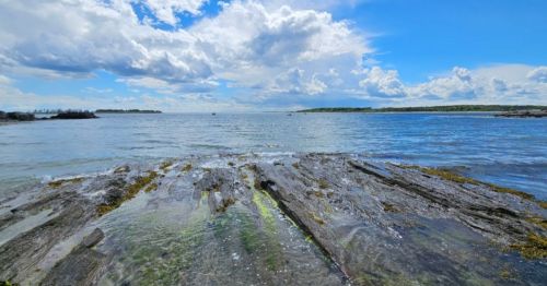 A serene coastal scene with rocky shorelines, clear water, and a bright blue sky filled with fluffy clouds.
